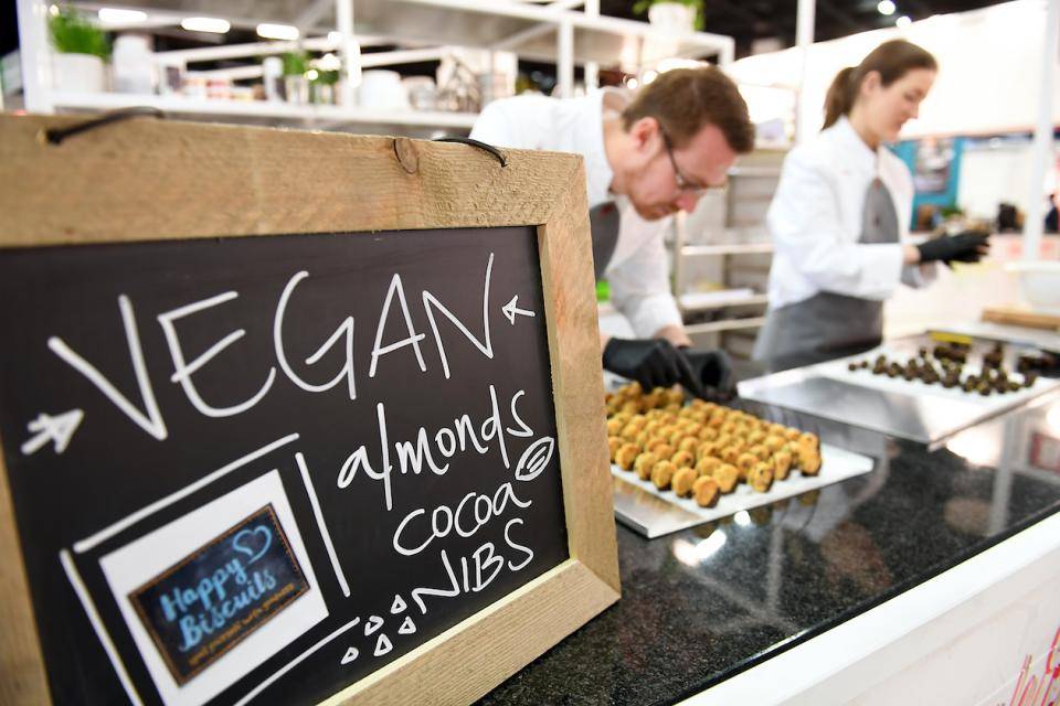 Chalkboard sign reading "Vegan" in foreground, chefs (blurred) working in background